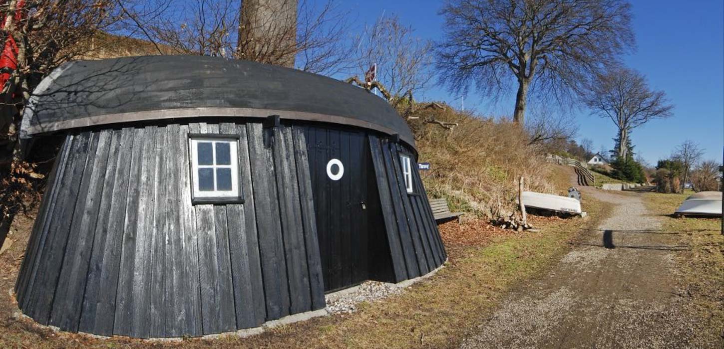 Sortmalet fiskerhytter i grønlige omgivelser. I baggrunden ses et par både, der er vendt på bunden i græsset.