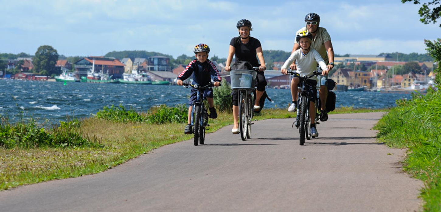 En familie på fire cykler sammen langs en sti ved kysten. Børnene fører an, og de voksne følger efter. I baggrunden ses det blå vand med en by helt i baggrunden. Det er en klar dag med solskin.