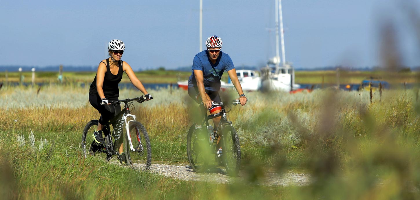 To cyklister på grussti i grønt landskab med to hvide både i baggrunden.