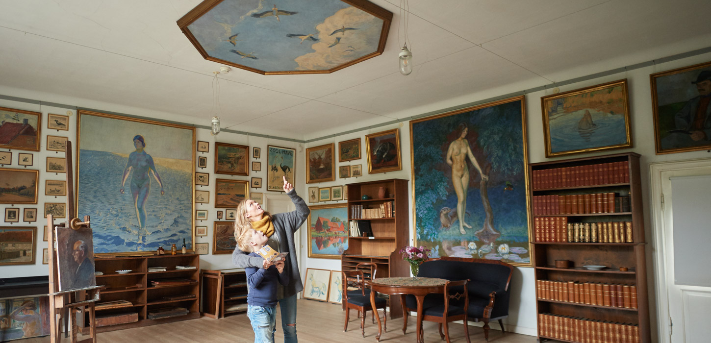 Mor og søn kigger på loftmaleri på Johannes Larsen Museet. De er omgivet af malerier lavet af Johannes Larsen