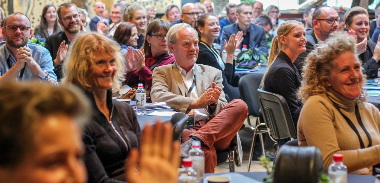 Rundt om runde blå borde sidder en masse mennesker. De kigger alle sammen i samme retning og sidder og klapper.