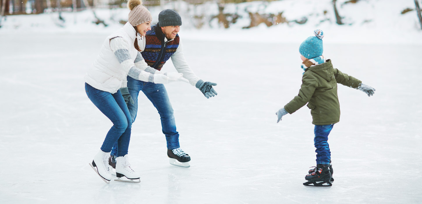 To voksne udstrækker armene mod et barn, som skøjter imod dem i et snedækket skovlandskab.