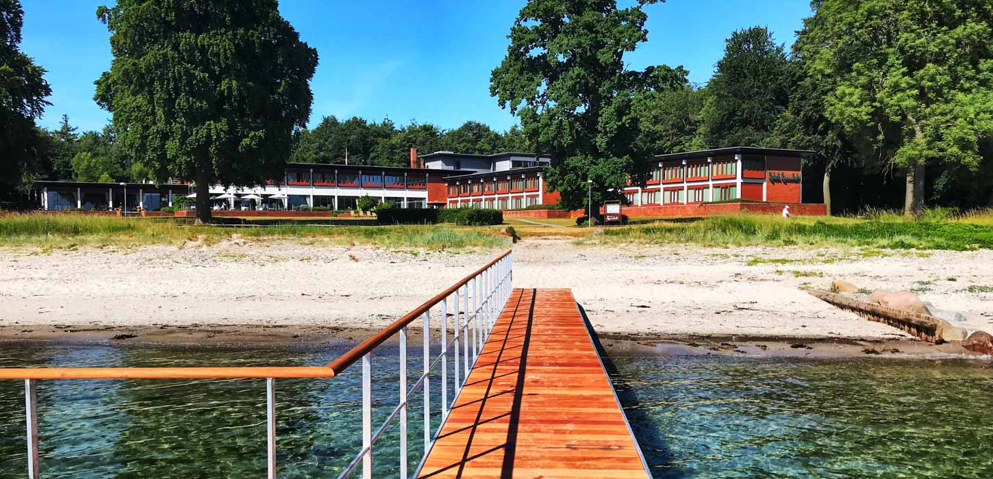 Udefra havet går en træ-badebro ind til sandstranden. I baggrunden ligger det røde Hotel Hesselet gemt bag træerne.