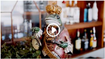 En frø af keramik i fokus i forgrunden. Den bærer en krone, rødt tøj og holder et lommeur. Der er indsat en hvid afspilningsknap i midten af billede