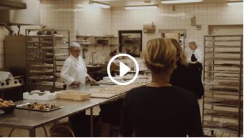 Køkkenet i OCC Odense Congress Center. To kokke står og laver mad, mens to kvinder kommer ind i køkkenet. Der er et videoikon i midten af billedet.