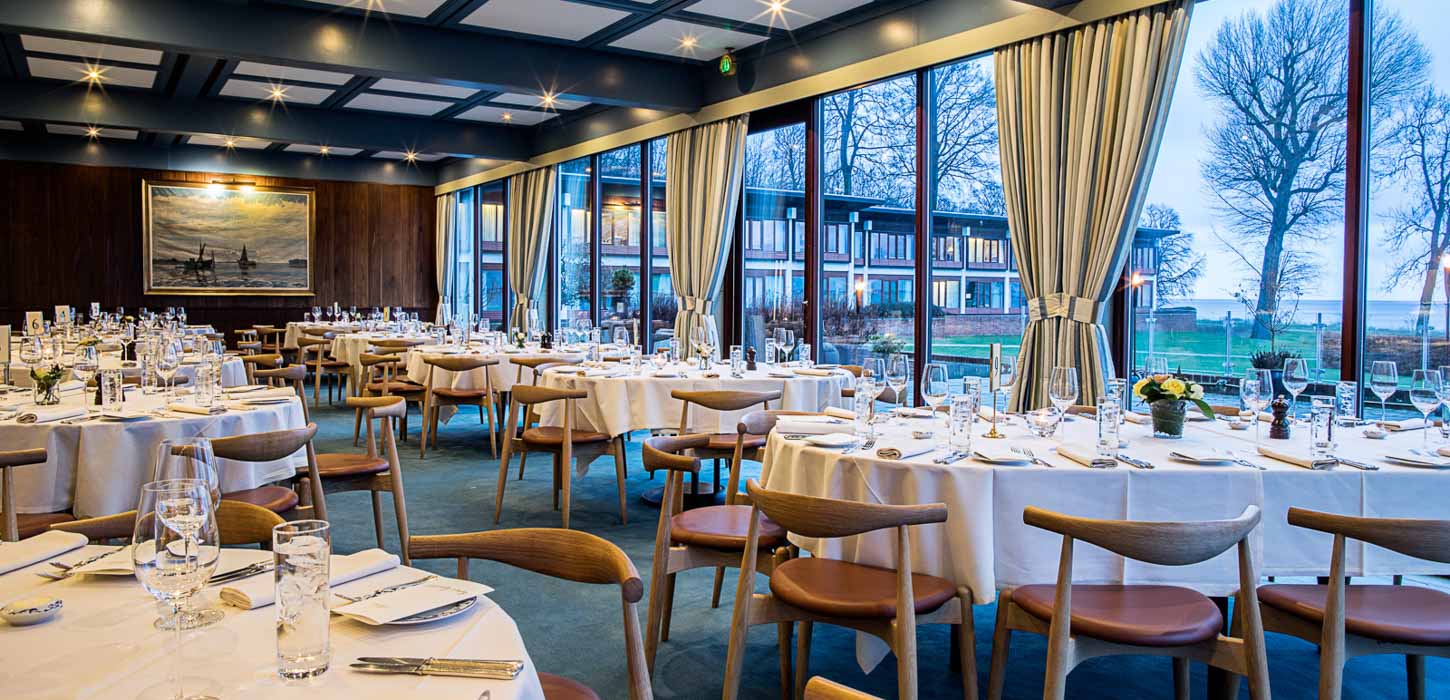 Et billede fra et stort mødelokale med store vinduer med udsigt udover vandet. I lokalet stor 7 runde borde med hvide duge og opdækning. Rundt om bordene står der træstole. 