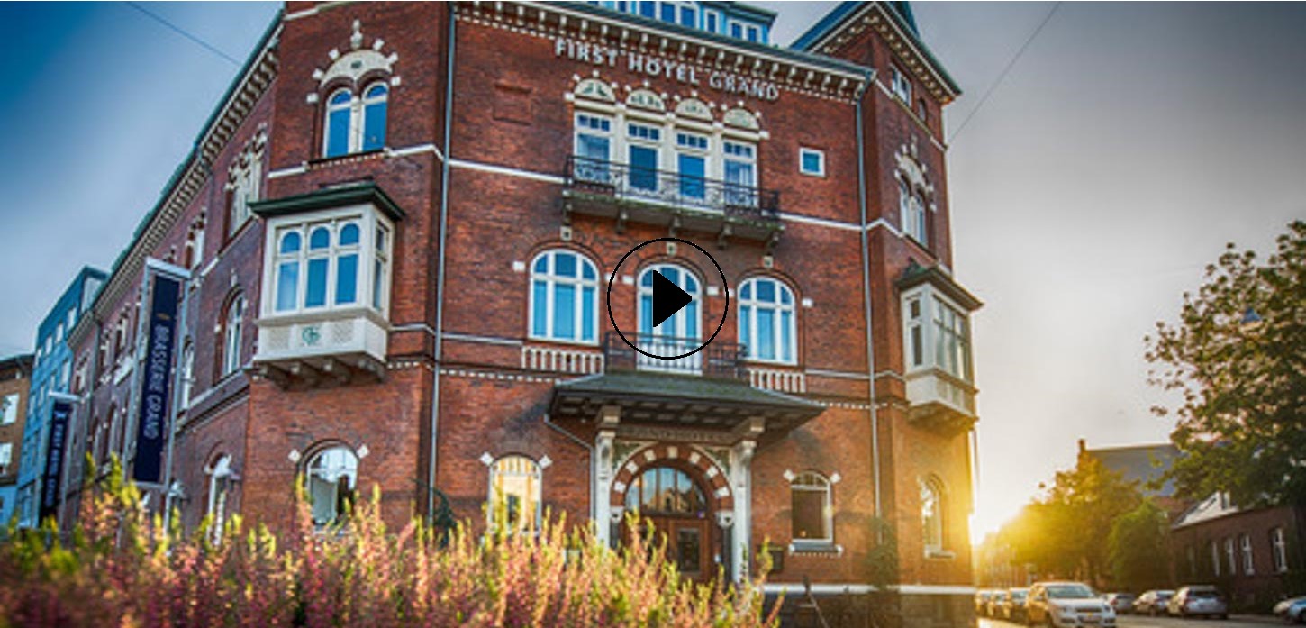 First Grand Hotel tårner mod himlen med en skarp solstråle i baggrunden. Der er en playknap på billedet, som der kan klikkes på for at afspille videoen.