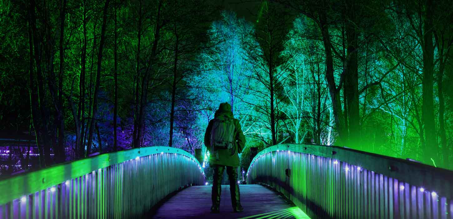 Midt på en bro står en mand i vintertøj. På hver side af ham er gelænderet på broen oplyst af blå og grønne lamper. I baggrunden ses halvoplyste træer.