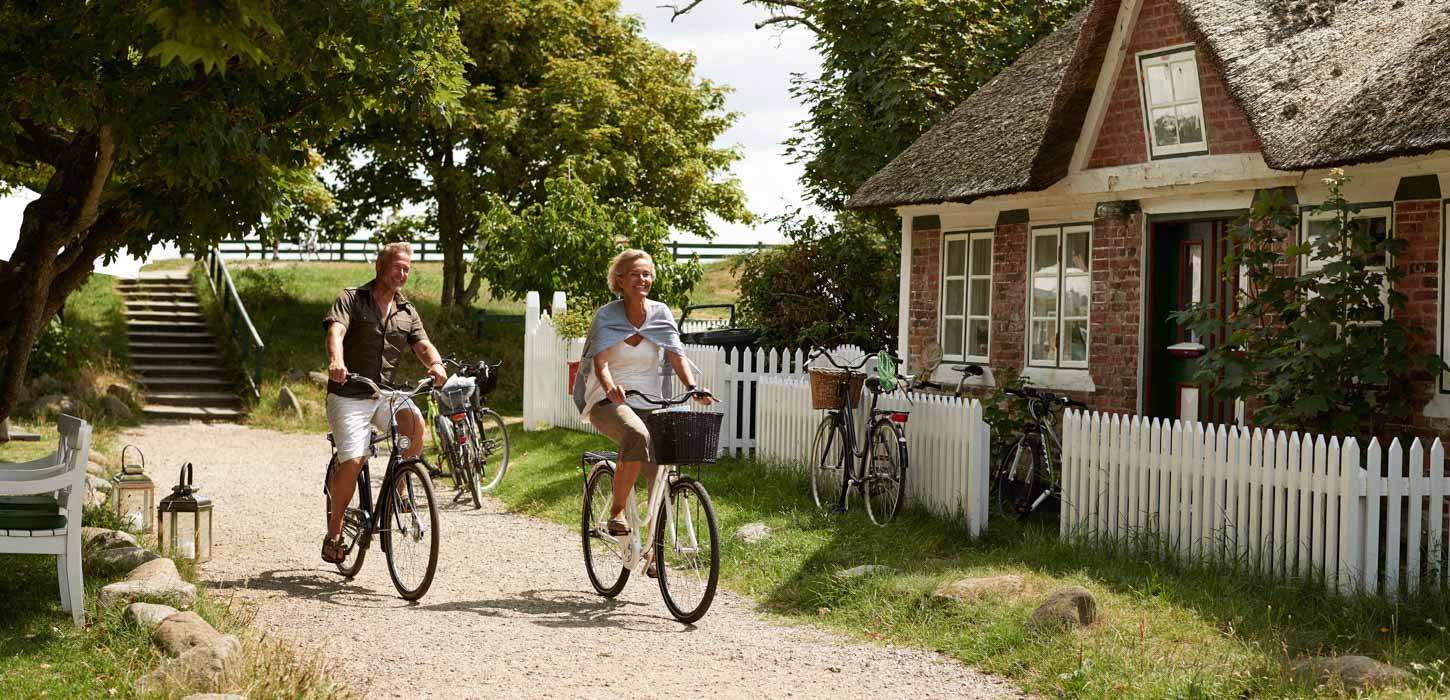 Et par cykler igennem idyllisk landskab med stråtagshus, hvidt stakit og grønne træer i baggrunden.