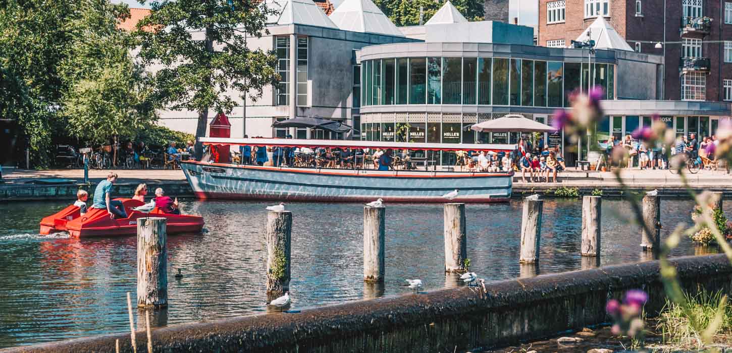 Odense Aafart Bådudlejning på en solskinsdag. En familie kommer sejlende i en vandcykel. Der sidder mange mennesker ude foran Nelle's i det gode vejr.  