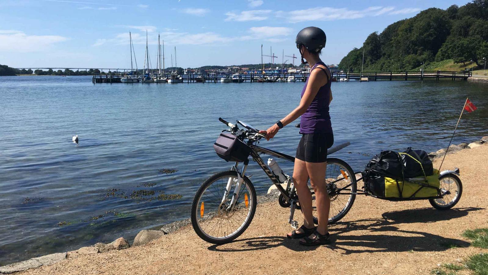 En kvinde står på en sti med sin cykel. cykelhjelm og cykeltrailer.  Hun kigger udover havet og havnen der er i baggrunden af billedet.