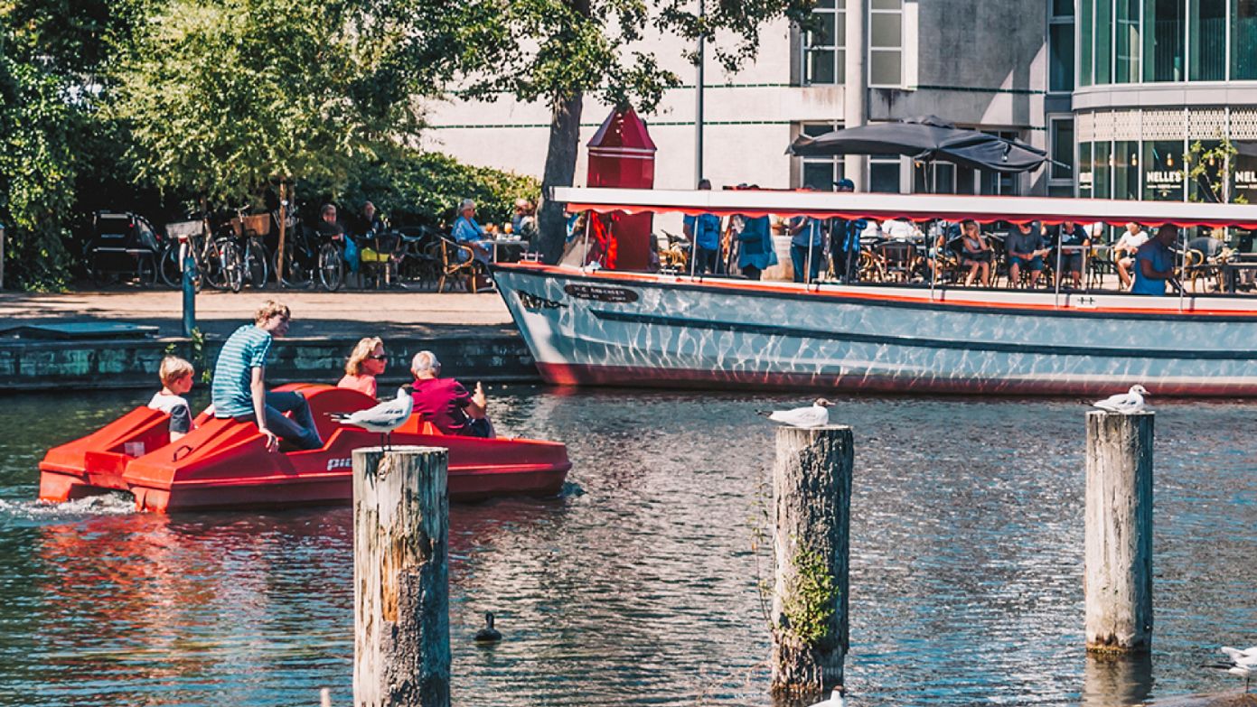 Odense Aafart Bådudlejning på en solskinsdag. En familie kommer sejlende i en vandcykel og mange mennesker sidder ude foran Nelle's i det gode vejr og kigger ud på åen..  