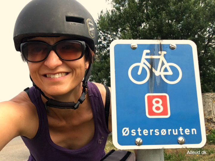 En kvinde i lilla tanktop, sorte solbriller og en sort cykelhjelm tager et selfie med et blå skilt. På skiltet er en hvid cykel og teksten "Østersøruten". i Bagrunden er en stort grantræ.