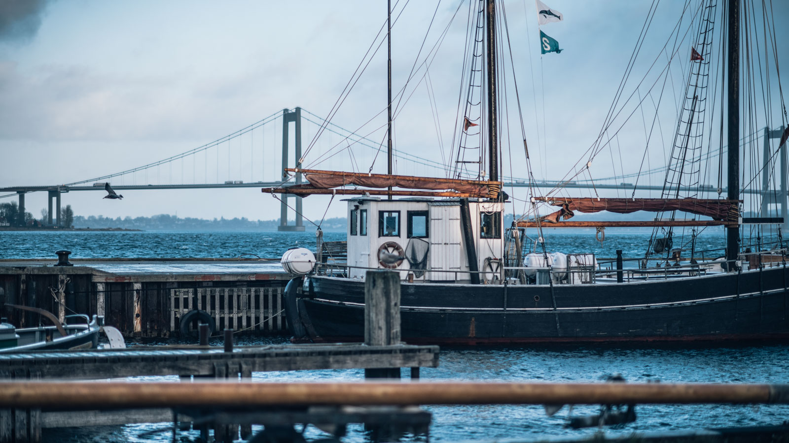Et gammelt blåt sejlskib ligger til kaj. I horisonten kan man se Lillebæltsbroen. 