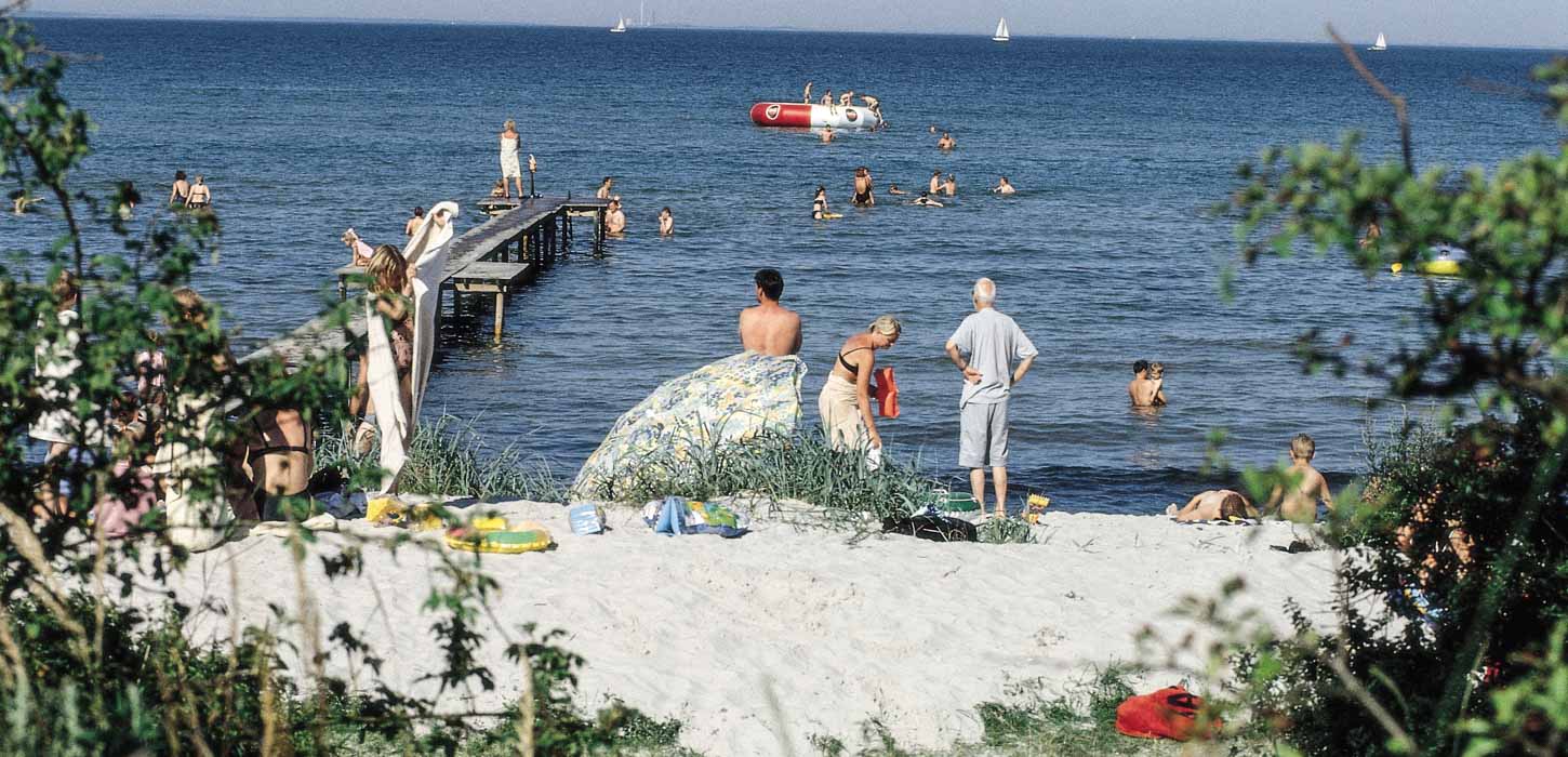 Strand med bølgende vand, badebro og mange badende gæster.