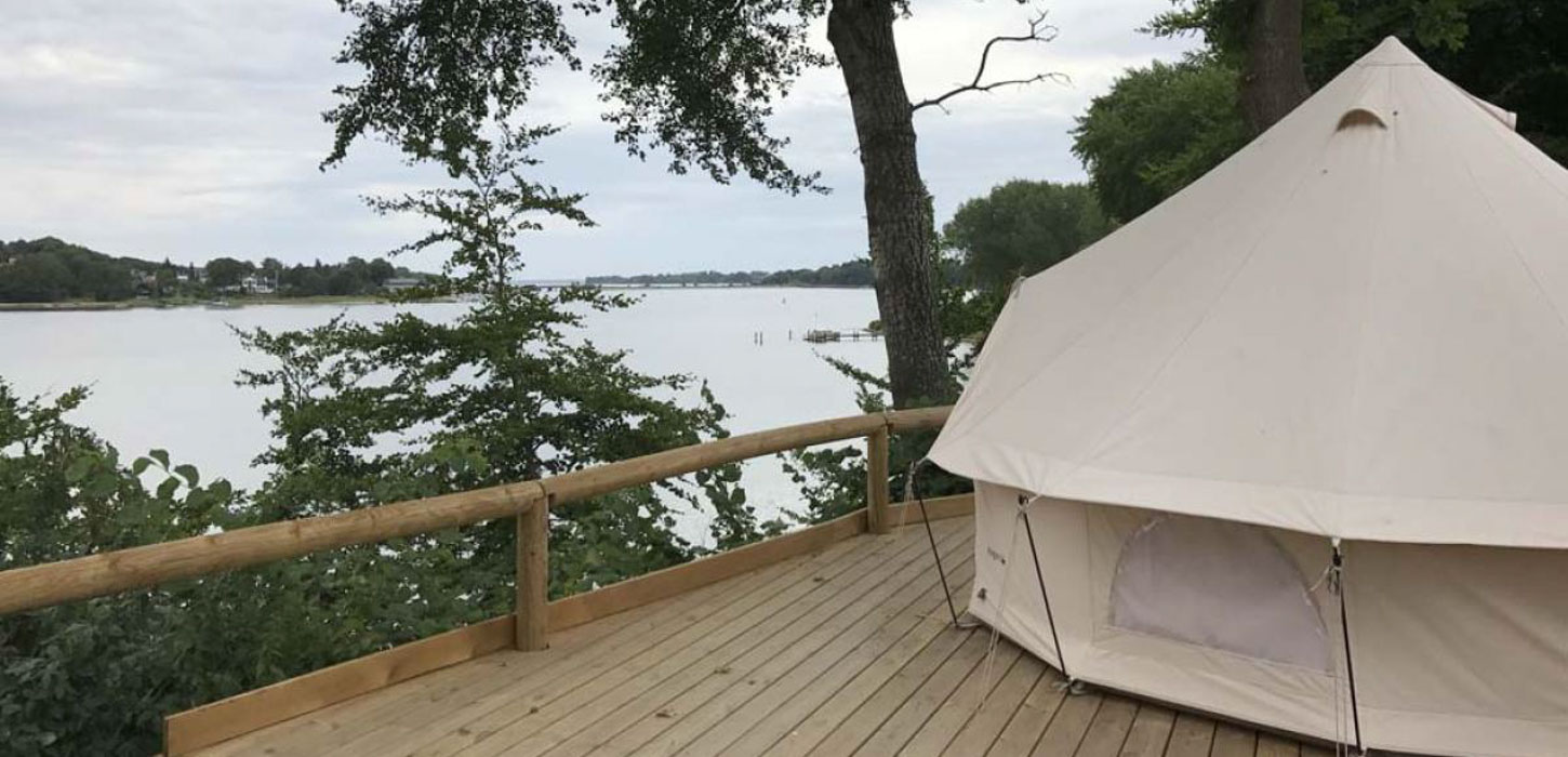 Lyst glampingtelt på platform af brædder med udsigt over vandet.