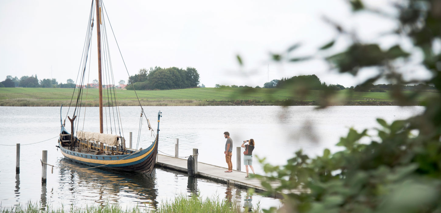 Det meste af billedet er består af vand som i horisonten mødes med græs. Midt i billedet ses en badebro, hvor der ligger et gammelt vikingeskib bundet fast. På badebroen står en kvinde og en mand. Kvinden fotograferer skibet.