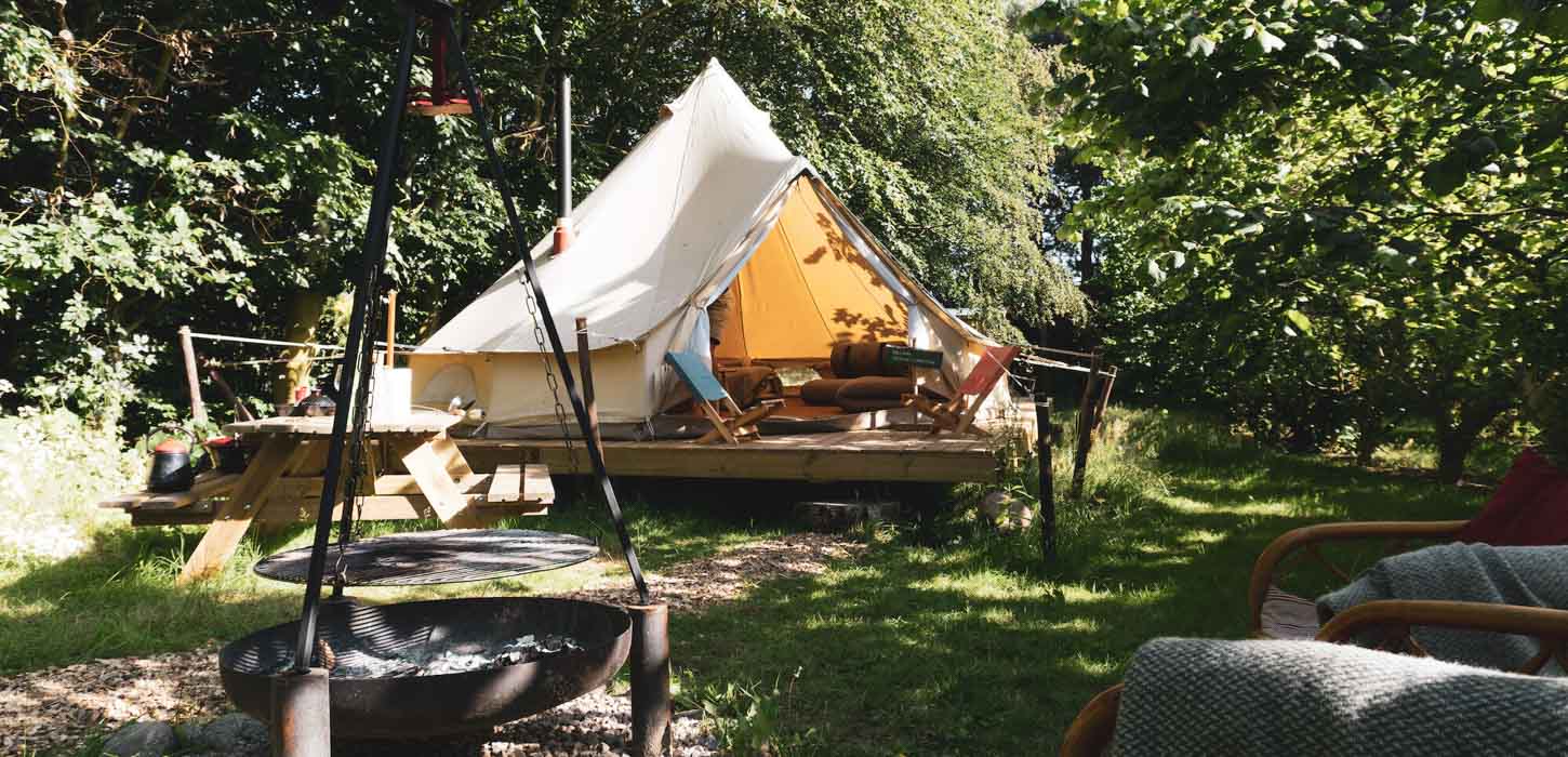 Åbent hvidt glampingtelt på træplatform med bålfad i forgrunden og grønne træer i baggrunden.