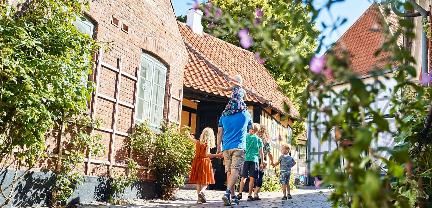 Familie med to voksne og tre børn går ned ad en brostensbelagt gade med bindingsværkshuse og stokroser.