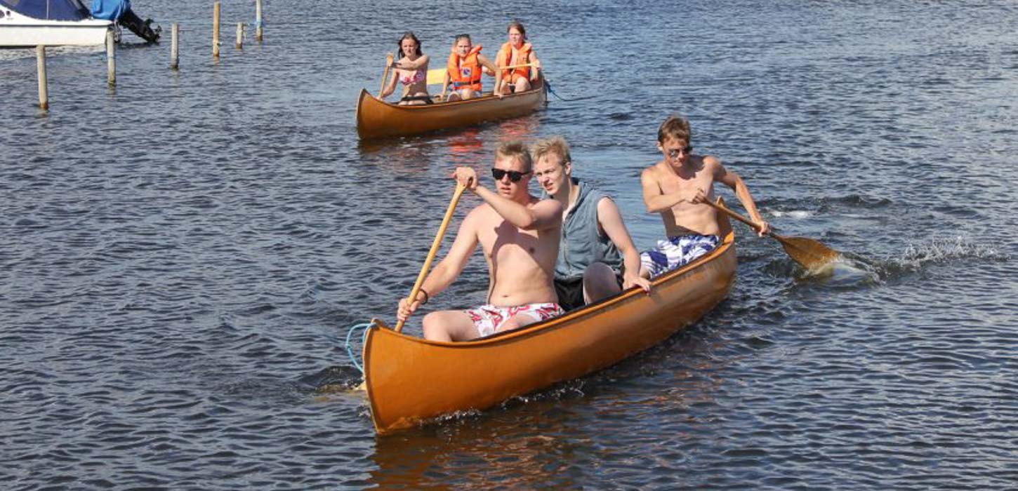 Det meste af billedet består af havvand. I venstre øverste hjørne ses bagenden af en lille båd og en par pæle der stikker op af vandet. På vandet sejler to kanoer forskudt af hinanden. I den bagerste sidder tre piger. To i redningsvest og en uden. I den forreste sidder tre unge mænd to i bar mave og en i en grå tanktop.
