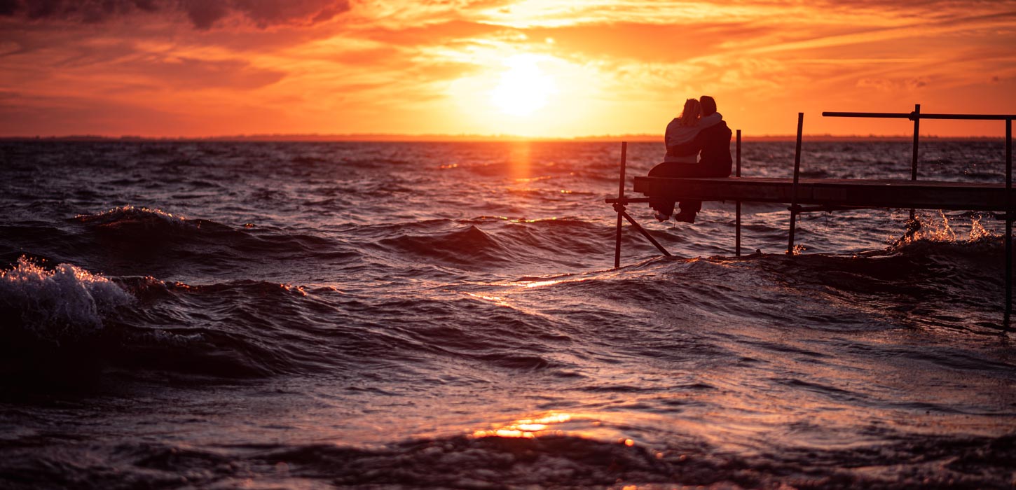 Et par sidder på en træbro over havet og iagttager en intens rød solnedgang.