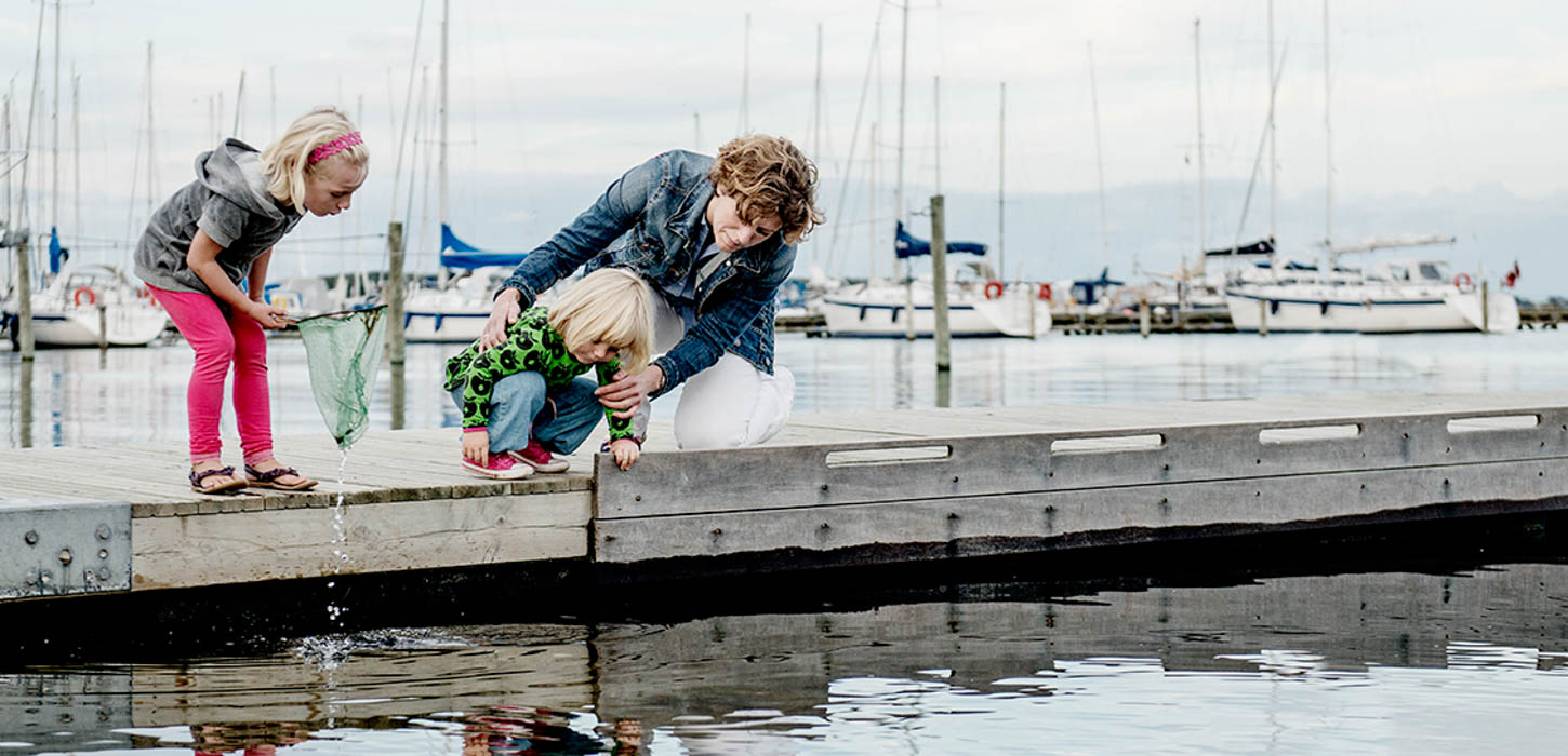 To børn og en voksen fisker med et fiskenet på en havnebro. Skibe i baggrunden. Den voksne holder fast i det mindste barn, der er nede i knæ og læner sig ud over kanten for at se.