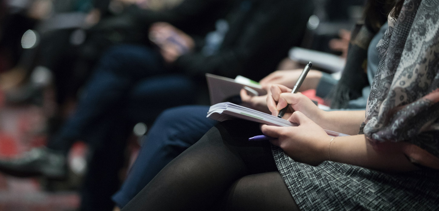 Et nærbilede af kvindehænder der skriver i en notes bog liggende på hendes krydsede ben. Man kan ikke se hendes hoved. I baggrunden kan man se at hun sidder på en række, hvor nogle mennesker gør det samme.
