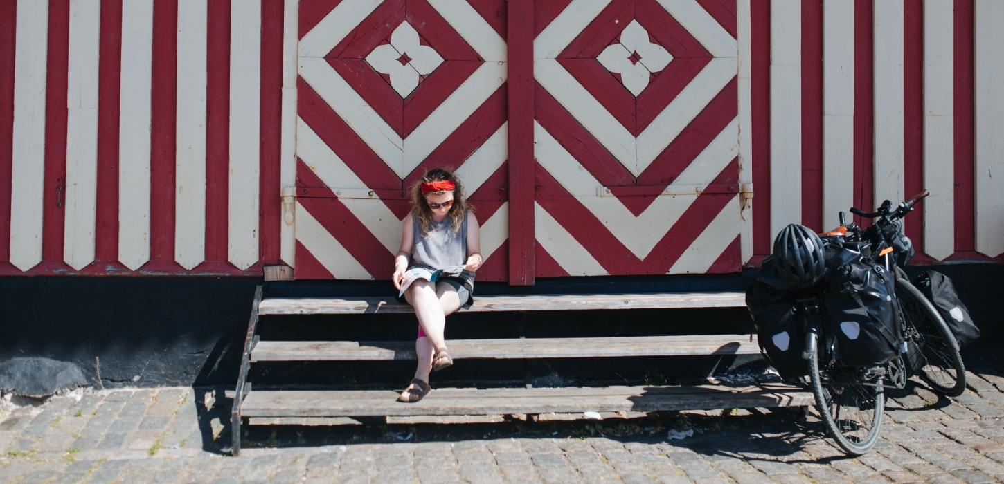 Kvinder sidder på trappen ved rødt og hvidt pakhus på havnen i Svendborg. Hun læser i et blad. Ved siden af hende står en cykel med oppakning