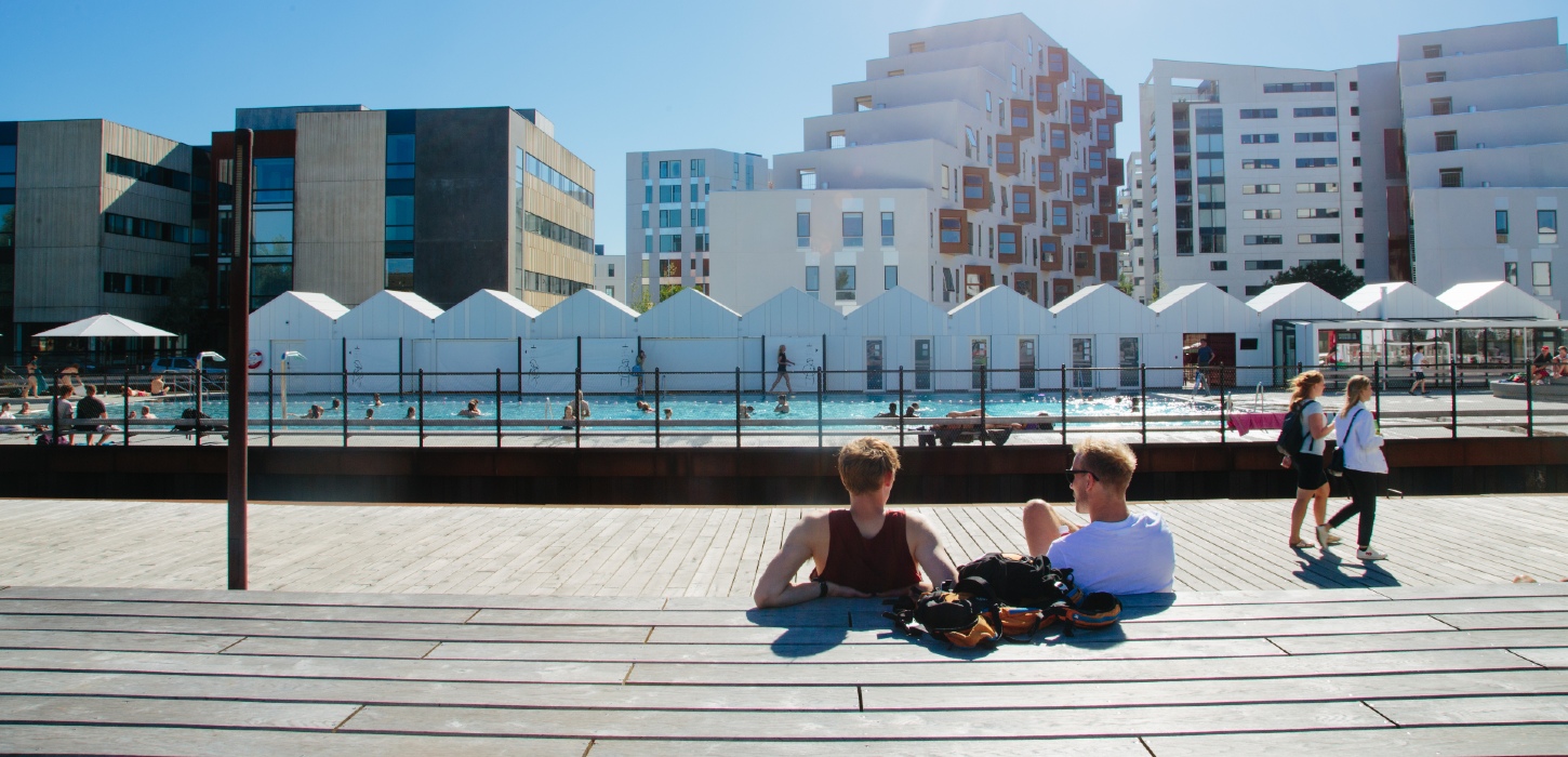 To drenge sidder med ryggen til ved havnen i Odense og kigger ud på havnebadet på en solskinsdag. To piger går en tur foran dem. 