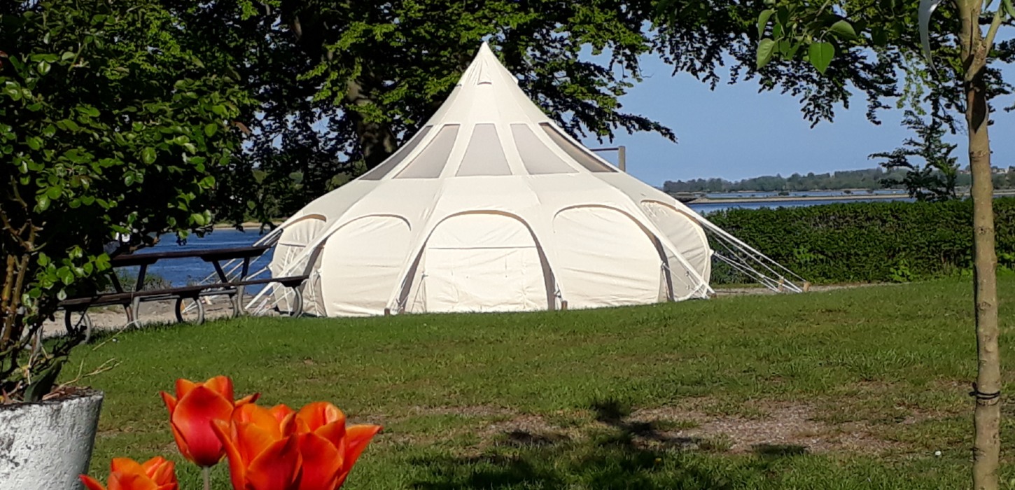 Stort hvidt glampingtelt på græsplæne i grønne omgivelser med blåt hav og blå himmel i baggerunden.
