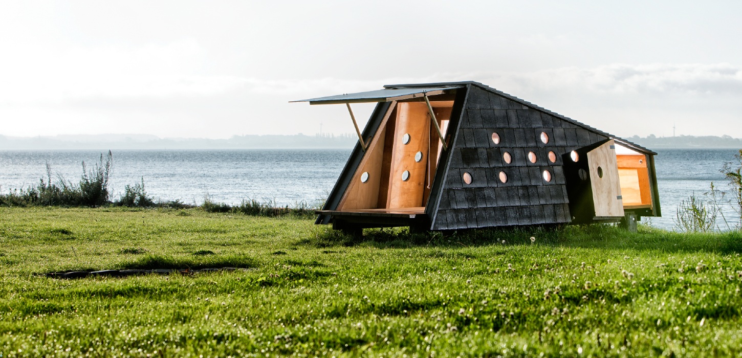 Shelter med åbne døre, der afslører et varmt interiør i træ. Det befinder sig i grønne omgivelser med udsigt over havet.