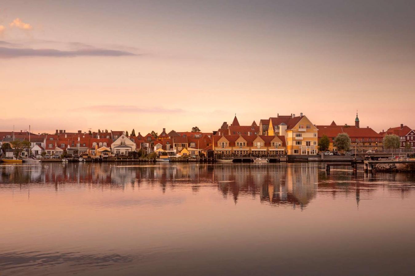 Kerteminde by set fra vandet. Solen er ved at gå nå så himlen er delvist lyserød. I forgrunden er der vand og i bagrunden ses Kerteminde by. Den består af en masse små bygninger i forskellige farver. De har dog alle røde tage.