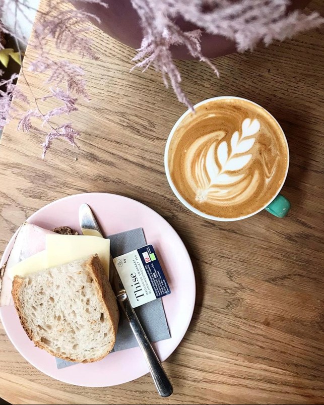 Kaffekop med lækker kaffe med flot mælkeskumsblad set oppefra sammen med tallerken med brød, ost, kniv, serviet og Thise smørbriket på træbord.