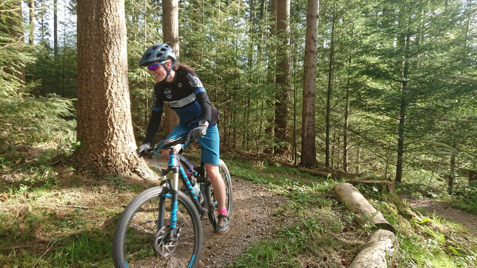 En kvinde i blåt cykeltøj cykler på et mountainbike-spor. Hun er på vej rundt i et sving og er omringet af skov.
