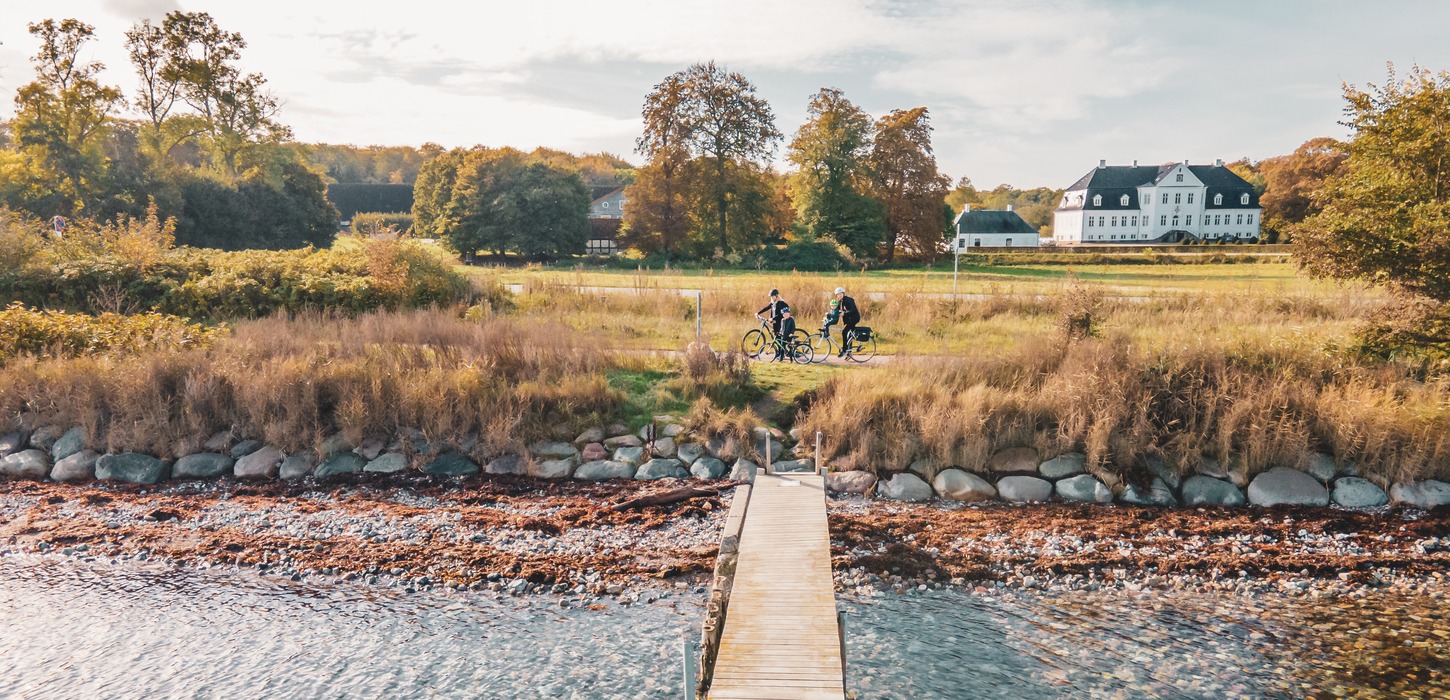 To cyklister gør holdet ved en badebro på strand med dige af store kampesten. I baggrunden en stor, hvid herregård og træer.