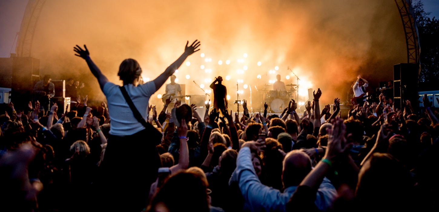 Foran en scene står et band oplyst af orange lys. Publikum står med begge hænder i vejret. En kvinde sidder på skulderen af en anden og hun har begge arme over hovedet. 