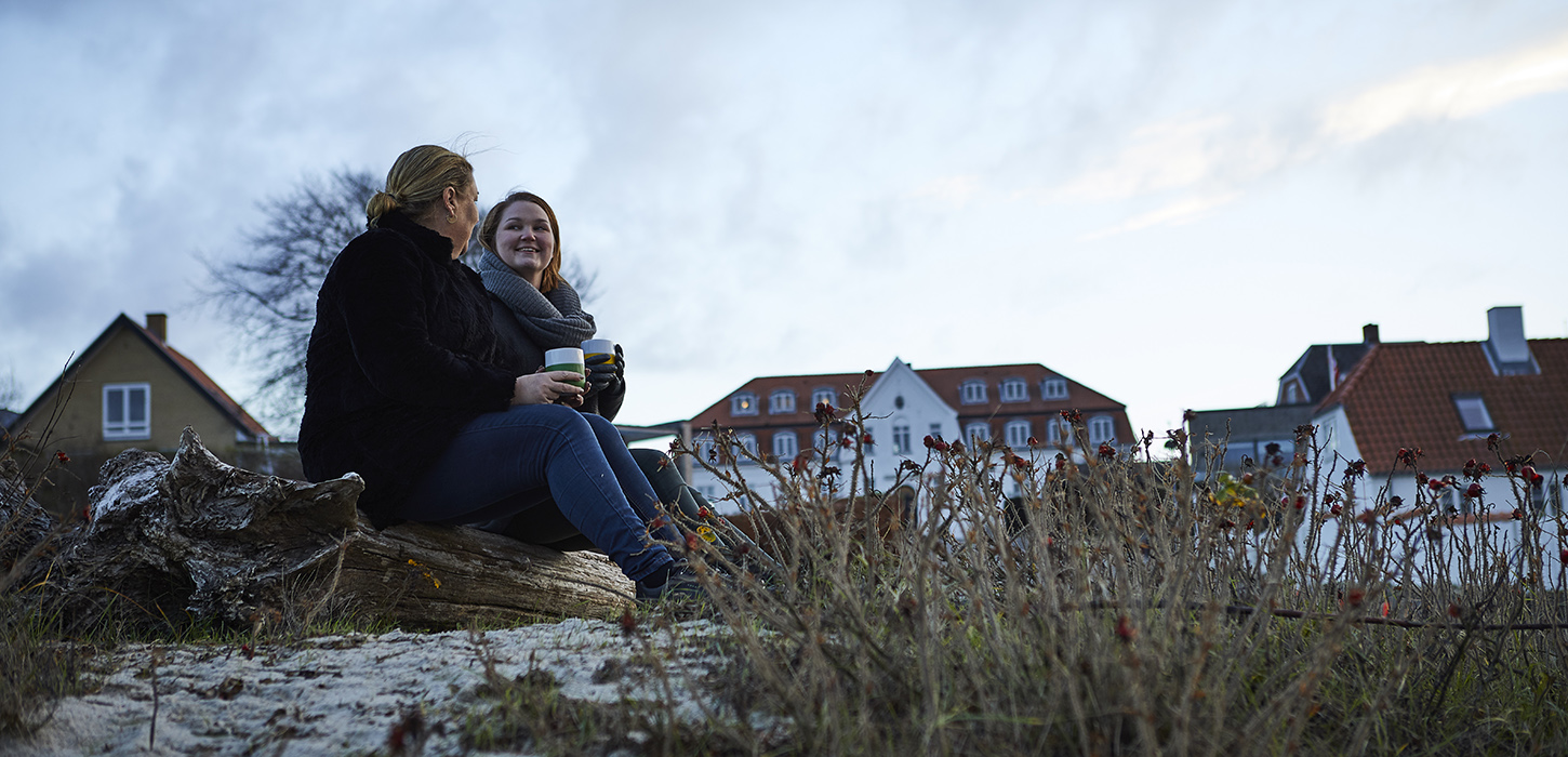To piger sidder trærester på jorden og drikker kaffe og bag dem er bygninger i baggrunden