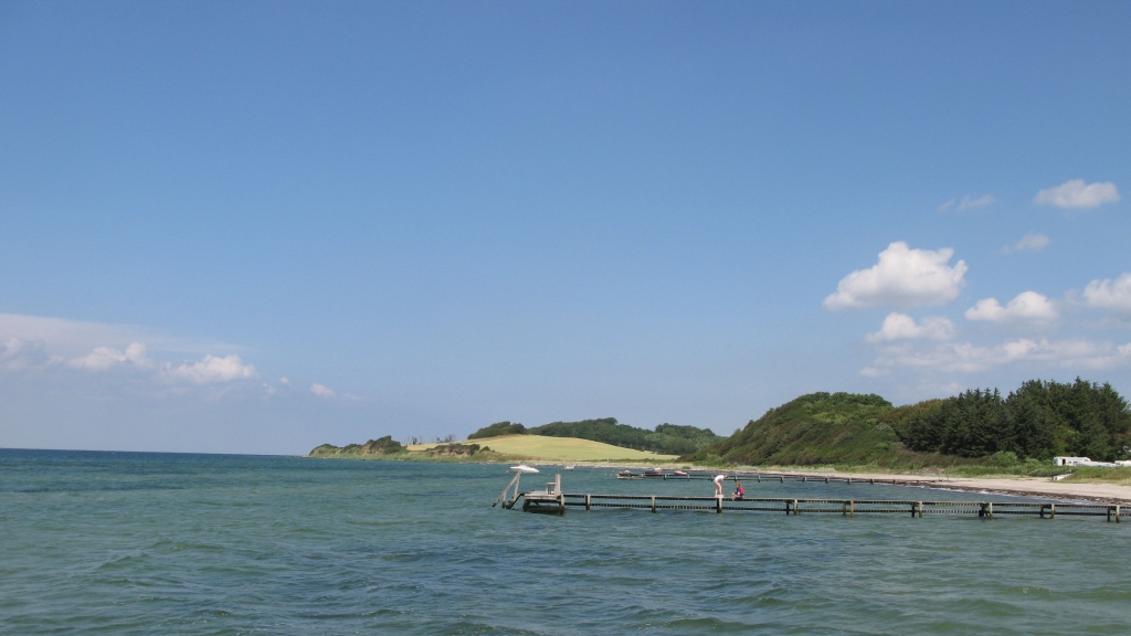 Kig over vand med badebro og strand og marker i baggrunden