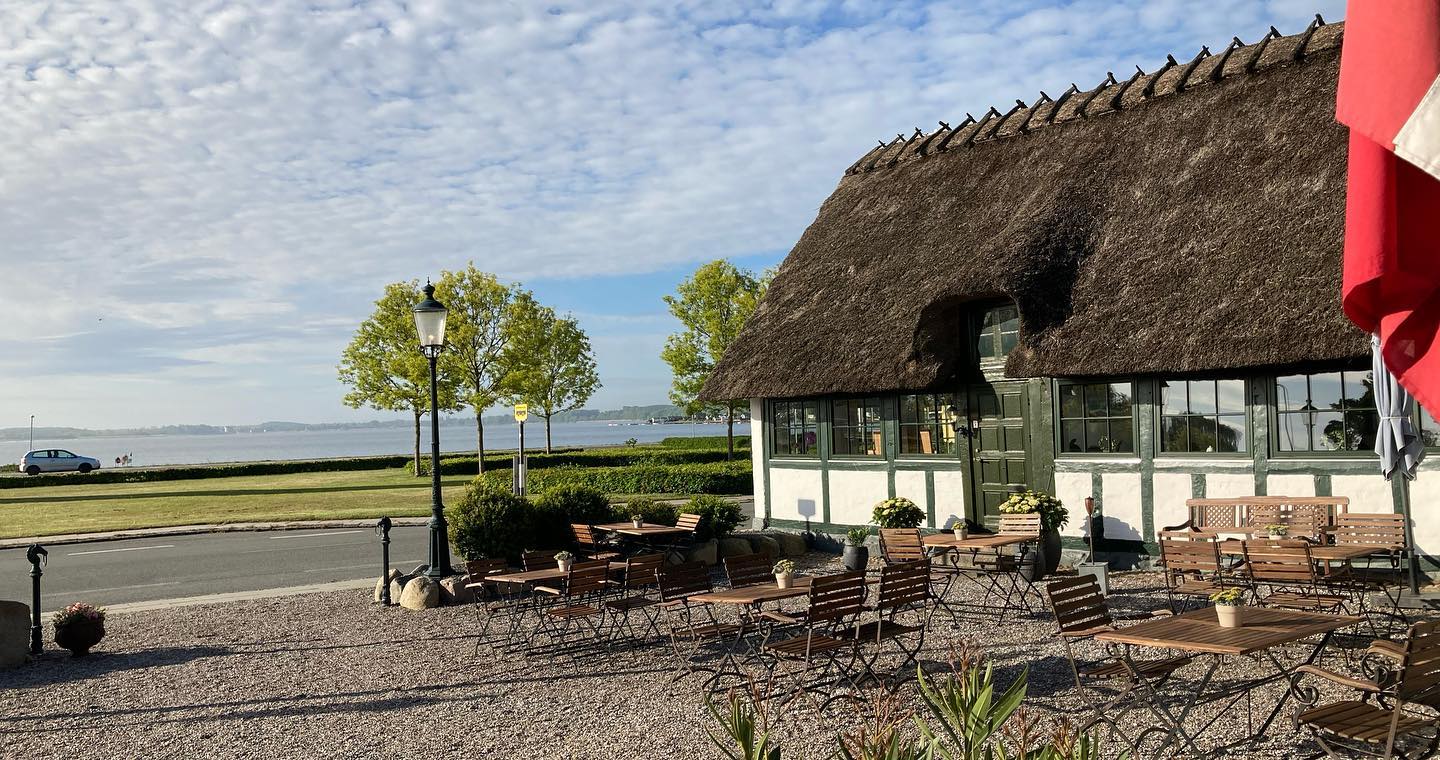 Længe af en hvid kro med stråtag og grønt bindingsværk, der ligger ud til vandet med en gårdsplads og borde og bænke.