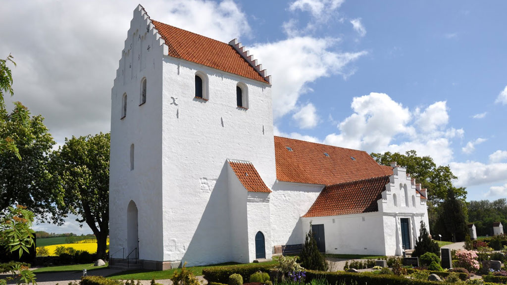 Foran en blå himmel står en hvis kirke med rødt tag. Den er omgivet af en kirkegård.