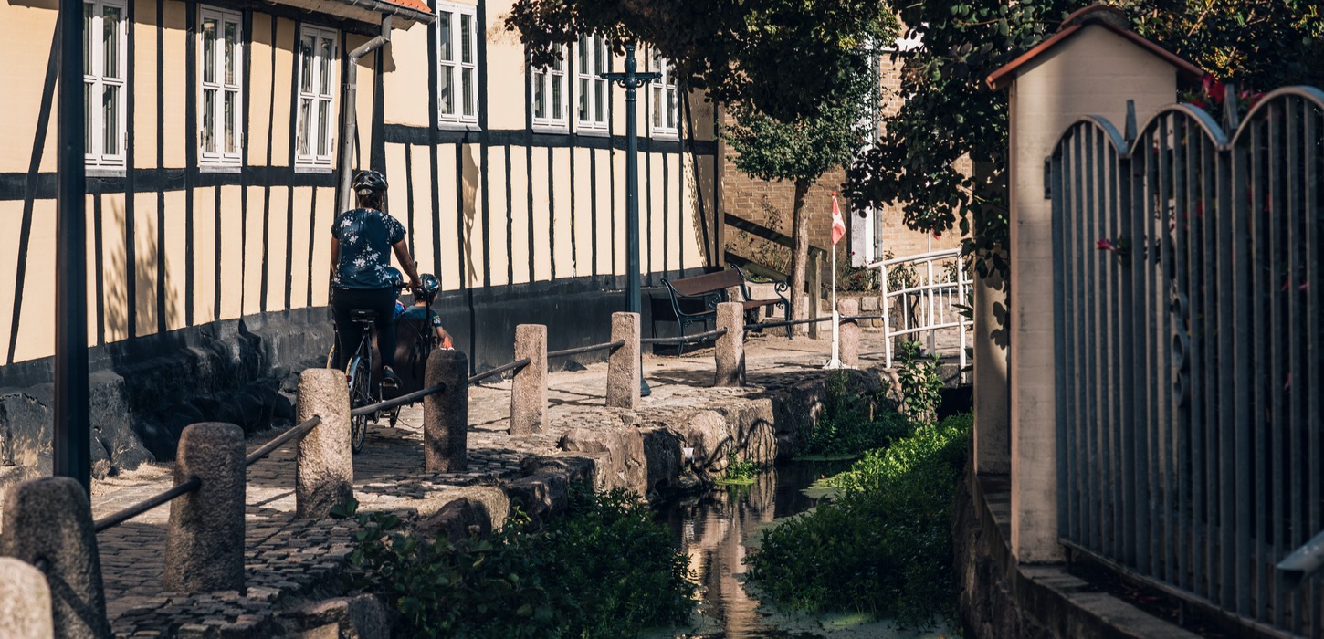 Cyklist trækker af sted med sin cykel på brostensbelagt sti langs med gult bindingsværkshus.