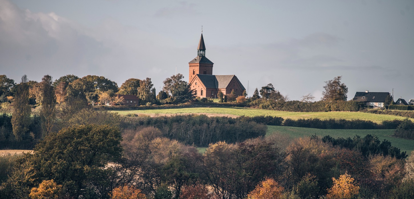 Bølgende landskab med krat, træer og grønne marker. I baggrunden troner en rød kirke frem.