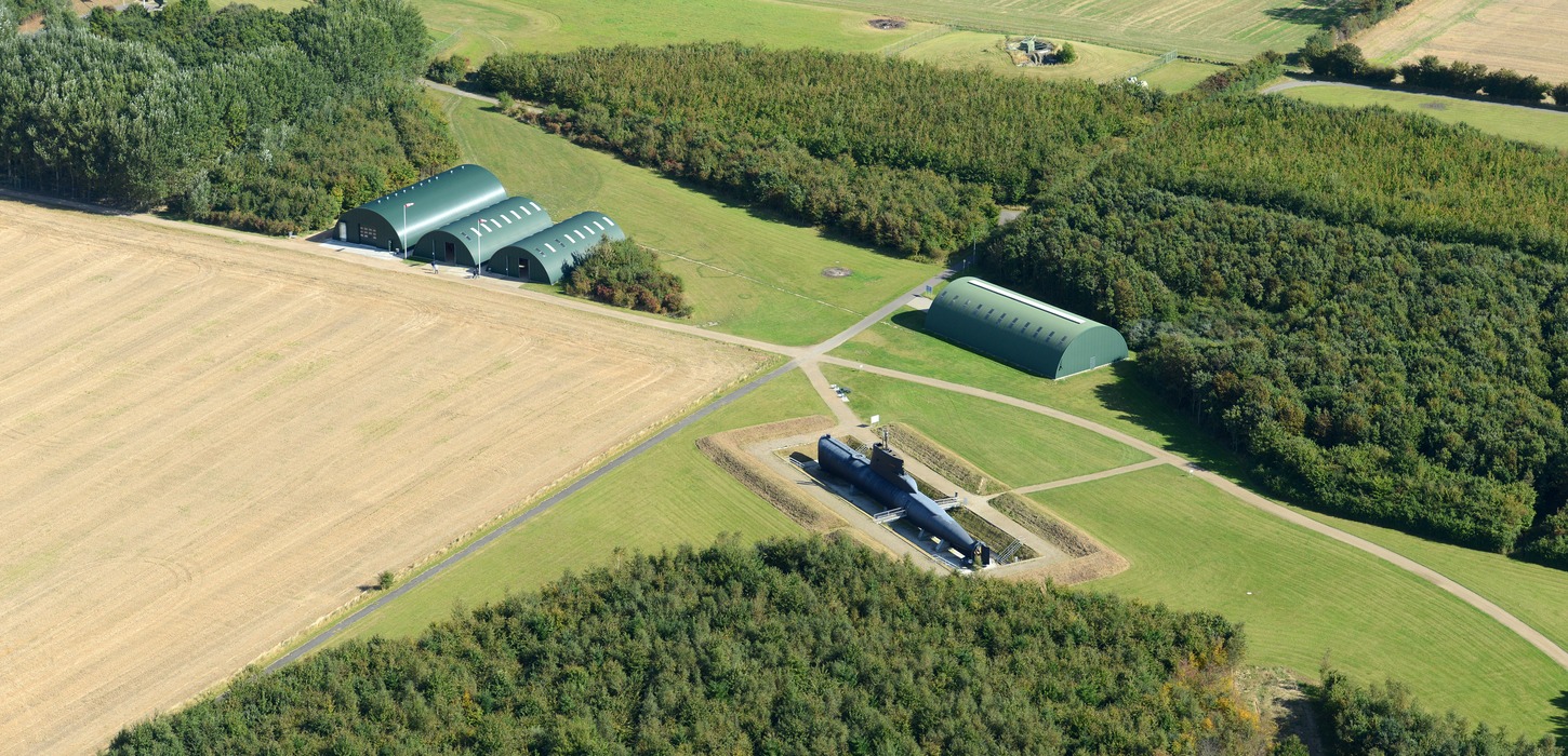 Dronebillede: I venstre side ses en lysbrun nyslået mark. I venstre side omringer skov fire grønne barakker, og en sort bygning.