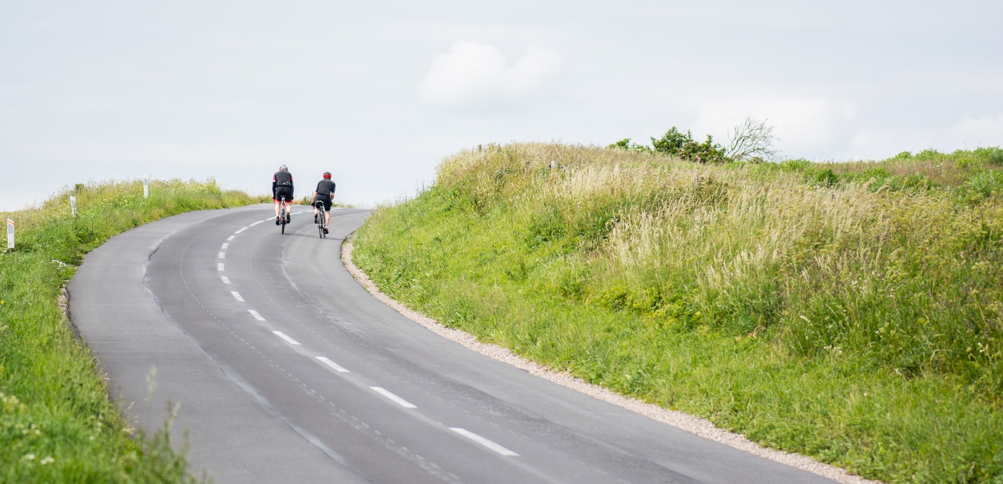 To cyklister på vej op ad bakke på landevej med grønt omkring. 