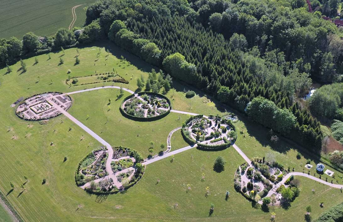 En dronebillede af en stor mark. Til højre for den er der skov og til venstre er endnu en mark adskilt af en række træer. Marken består af fem små runde haver. De består af buske og træer. Imellem af haverne går en sti. 