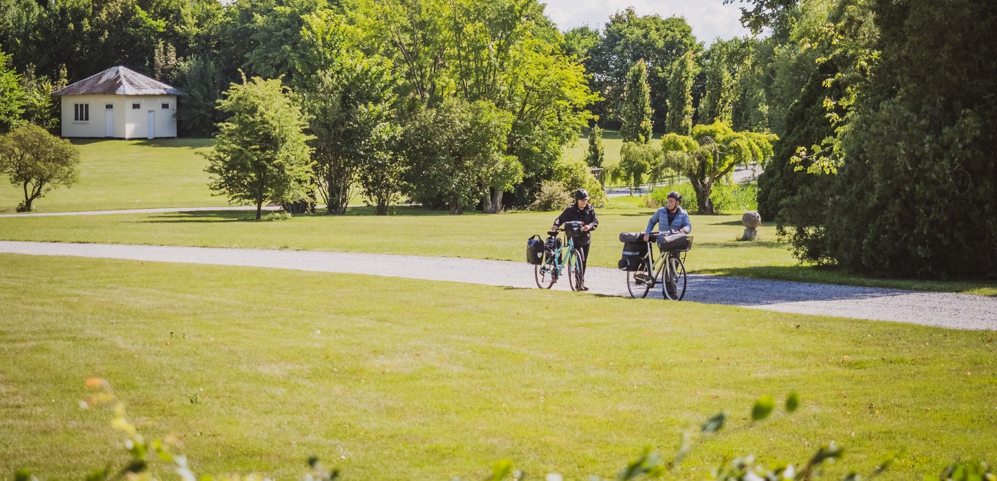 En par med hver deres cykel med opbakning går på en grussti. Foran stien er der græsplæne mens der bagved er græs og træer. 