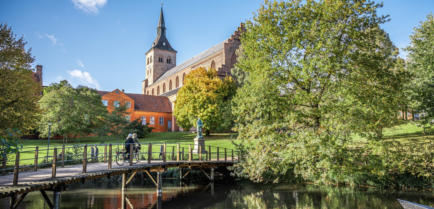 En idyllisk grøn park med en stor kirke med et spidst tårn i baggrunden. Kirken, der har en klassisk arkitektonisk stil, er omgivet af frodige træer i fuldt løv. Foran kirken er der en lille bro over en rolig å, hvor en person krydser på cykel. Himlen er klar og blå.