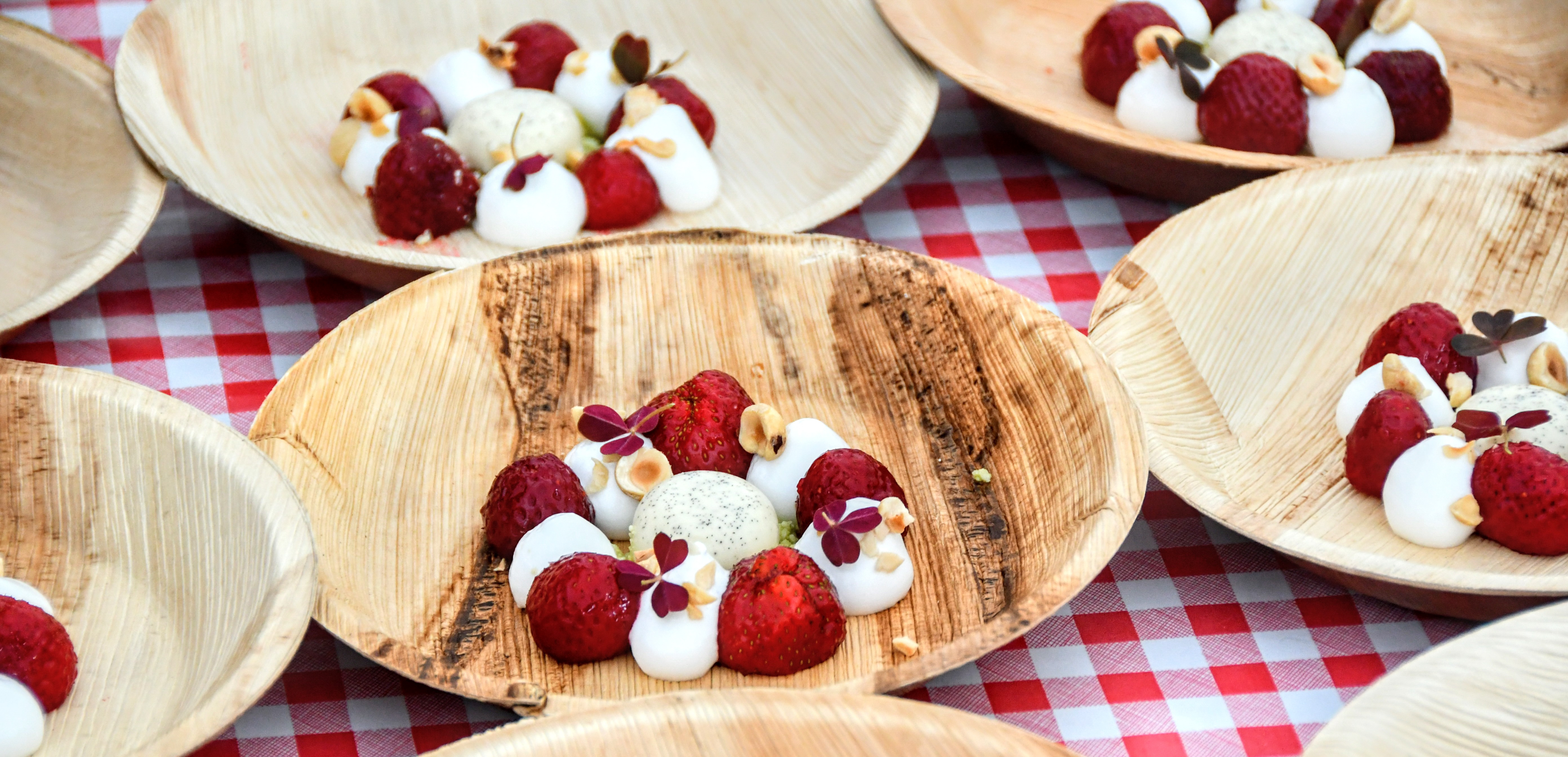 På en rød og hvid ternet dug står otte bæredygtige paptallerkner. På tallerknerne er en flot anrettet dessert bestående af jordbær, hvidt skum og en iskugle i midten. 
