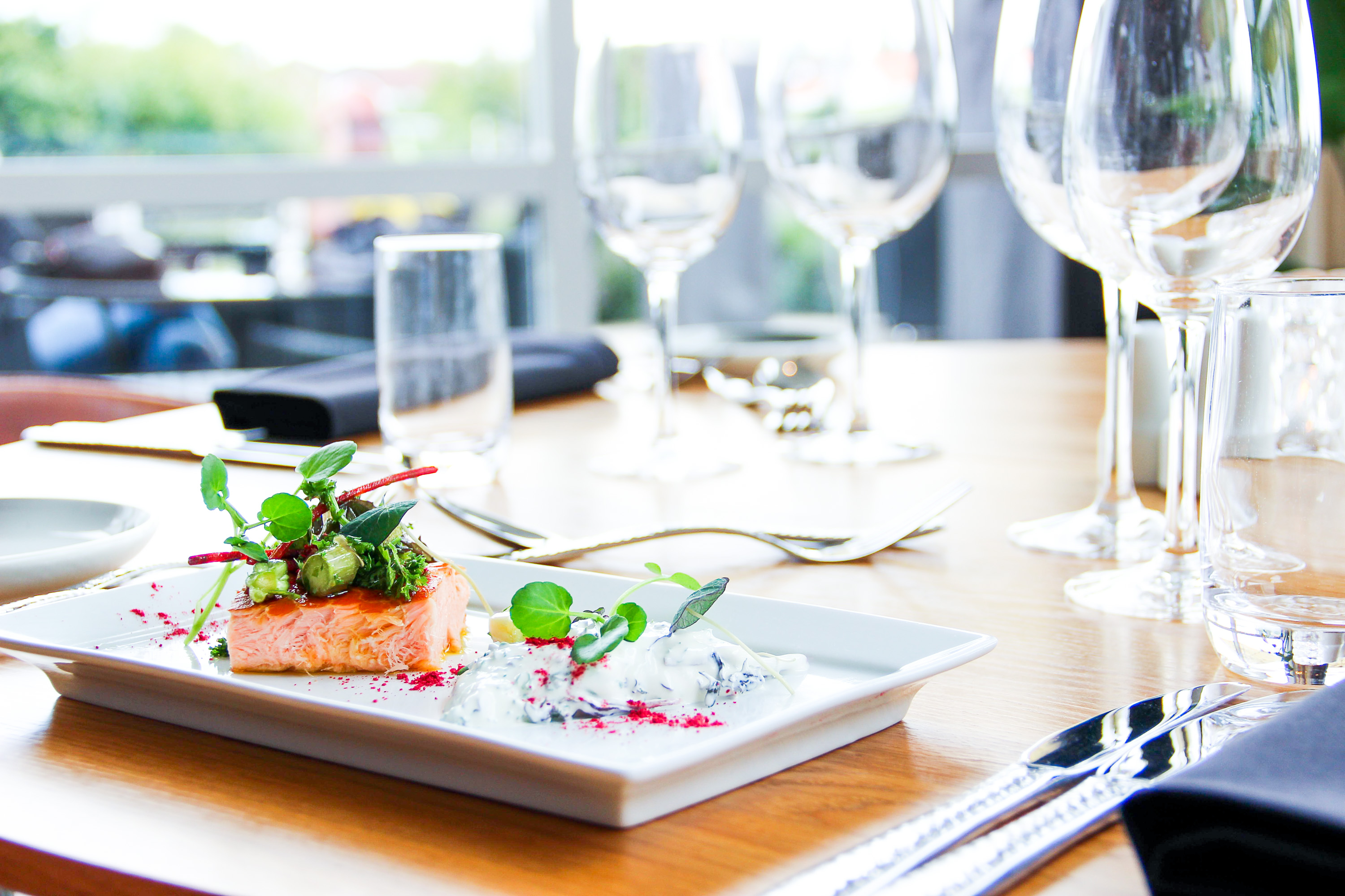 På en firkantet hvid tallerken ligger et stykke laks med grønt på toppen. Ved siden af ligger en hvid dressing som også er pyntet med ærteskud. I baggrunden ses slørede vinglas. 