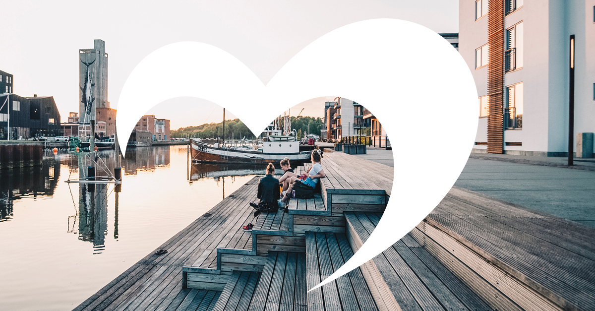 Stemningsbillede fra Odense havn en aften med solnedgang. Mennesker sidder på en træplatform ud til vandet og kigger på skibene. I forgrunden kan man se et hvidt Destination Fyn logo.